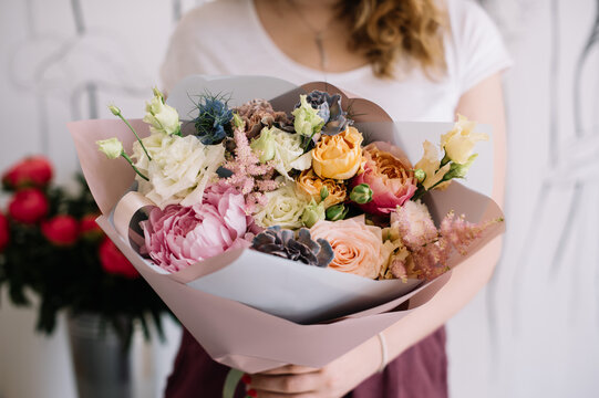 Very nice young woman holding big and beautiful bouquet of fresh peony, roses, eustoma, astilbe, carnations flowers in tender pink, yellow and blue colors, cropped photo, bouquet close up