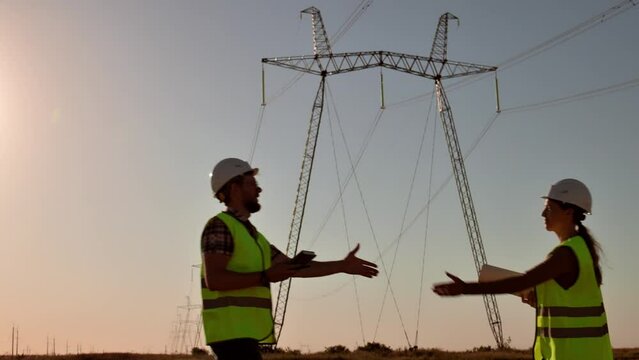 Two electrical engineers meet and shake hands against the backdrop of high-voltage electrical tower at sunset. Meeting of workers near power grids. Teamwork, signing contract, businessmen Slow motion