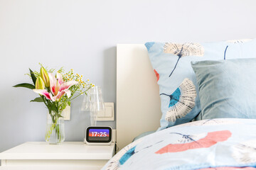 Beautiful bouquet of flowers with a blossoming pink lily in a glass vase decorates elegant and modern bedroom design with light blue colour theme during sunset, 17:25pm on digital clock