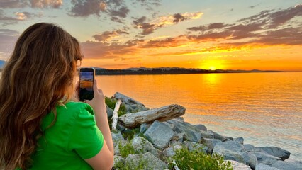 a teenage girl in a Green T-shirt stands with her back to the camera and shoots a beautiful sunset...