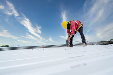 A Roofer working on roof structure of building on construction site, Roofer using electric drill nail gun and installing white Metal Sheet on top new roof. Safety body construction © tong2530