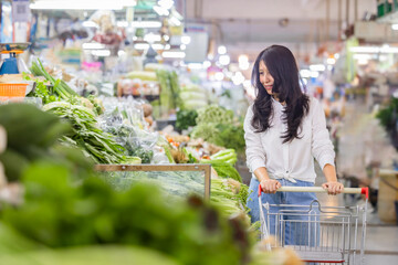 Organic food market. Middle age asian housewife pushing shopping cart walking near vegetables...