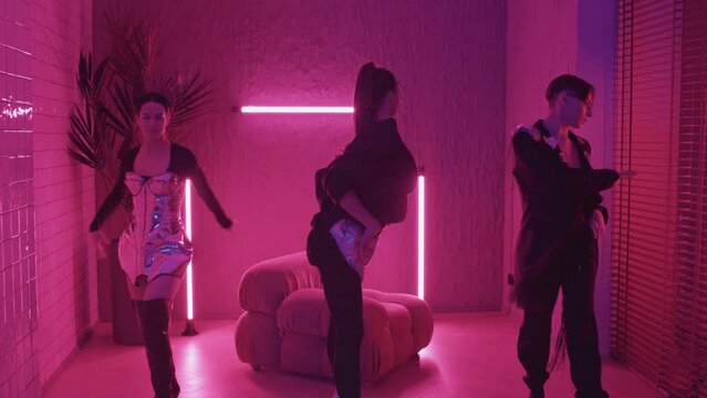 Tracking out of young stylish girls and guy in eccentric outfits dancing vogue in studio with pink neon lights