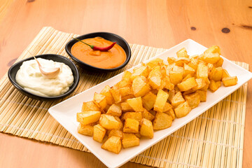 Fried diced potatoes on a white plate next to two bowls of aioli sauce and hot sauce.