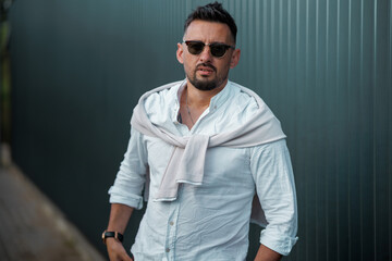 Fototapeta premium Fashion model handsome young man with trendy hairstyle in sunglasses in white shirt in light jeans posing near vintage wall on street on sunny day. Attractive american guy on walk. Copy space.