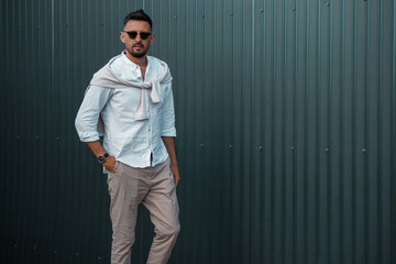 Fashion model handsome young man with trendy hairstyle in sunglasses in white shirt in light jeans posing near vintage wall on street on sunny day. Attractive american guy on walk. Copy space.