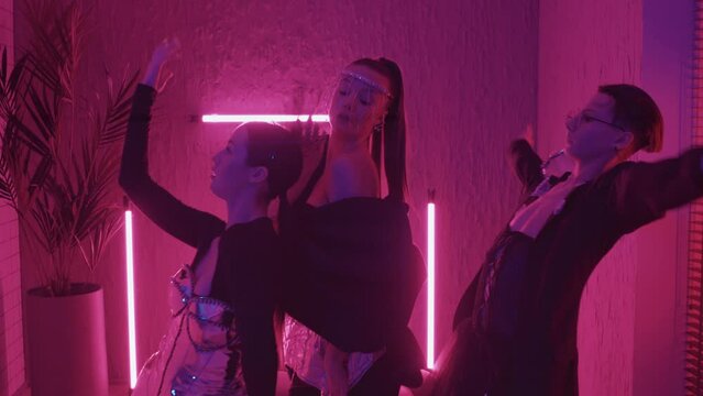 Slow motion portrait of young stylish girls and guy in eccentric outfits dancing vogue in studio with pink neon lights, looking on camera