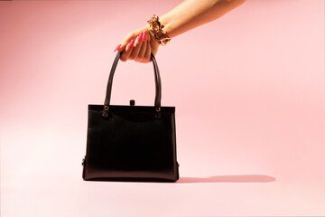 Female hand with golden bracelet holding black leather bag isolated on pink background