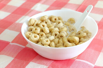 Healthy morning oat cereal. Cheerios in bowl of milk with spoon. Low sugar and high fibre breakfast. Isolated on red and white square patterned tablecloth.
