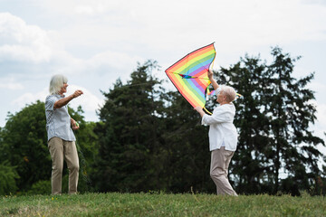 senior woman in casual clothes playing with kite near happy husband on green hill.