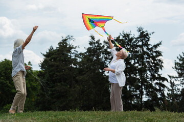 happy senior woman in casual clothes playing with kite near husband on green hill.