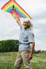 happy senior man in casual clothes holding kite and standing on green hill.