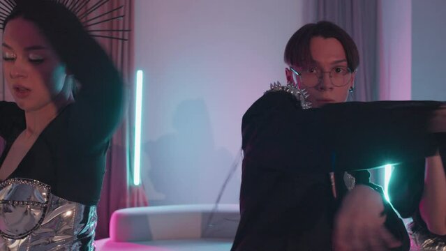 Tracking left medium of beautiful girls and guy in futuristic outfits dancing vogue in modern apartment in neon lights