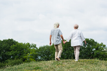 full length of senior couple in glasses holding hands and walking on green hill in summer.