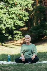 happy senior woman with grey hair sitting with crossed legs on fitness mat.