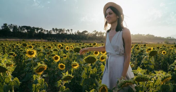 Happy romantic lady walk on sunflower meadow touching flowers and smile on summer vacation. Joyful female tourist in dress walk through plants on journey. Young adult woman with hat on sunny day