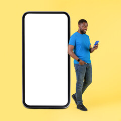 African Man Texting On Smartphone Near Large Cellphone, Yellow Background