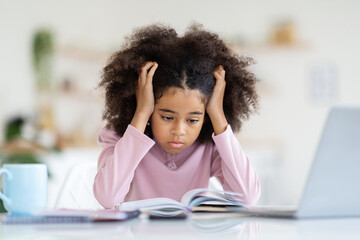Stressed black school girl sitting at table, looking at notepad