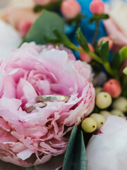 Wedding rings lie in a pink peony bud. In the background are flowers, petals, and berries in a bouquet.