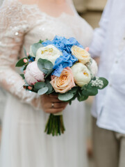 The bride stands and holds a wedding bouquet close-up. Bouquet of blue hydrangea, cream roses, white and pink peonies, eucalyptus leaves, and berries.