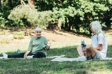 cheerful senior woman sitting on fitness mat and pointing at husband with sports bottle.