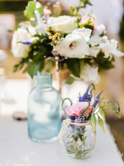 A field bouquet of rose and lavender stands in a glass vase on a white table. Floral decor in the background.