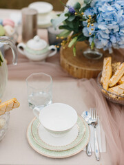 Table setting for tea. Porcelain cup on a saucer with a green border, cutlery, cookies in a crystal vase, and a bouquet of blue hydrangeas.