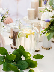 Fototapeta na wymiar Lemonade with lemon and lime slices in a glass jug stands on a white table. green leaves in the foreground, candles in the background.