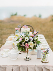 The decor of the wedding banquet table on the sea coast. Bouquet of white and pink flowers in a candlestick, wildflowers, teapot, and cutlery. The sea is in the background.