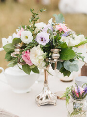 Floral decoration of the banquet table. A bouquet of white and lilac eustoma, pink roses, twigs, and leaves stands in a copper candlestick. In the foreground is a bouquet of wildflowers in a jar.