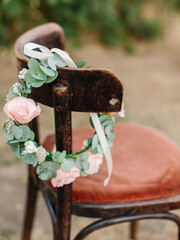 A wreath of pink rose and eucalyptus leaves with a white ribbon hang on the back of an old wooden chair.