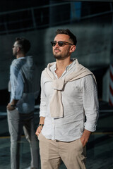 Brutal bearded man hipster in fashionable summer white clothes in sunglasses in stylish white sneakers stands on a sunny day near a glass building. Nice guy. Trendy menswear. Street style