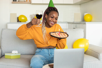 Online birthday. Happy black woman celebrating b-day showing pie with candle and glass of wine to...