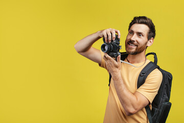 Positive middle aged man with backpack taking picture on professional DSLR camera over yellow background, free space