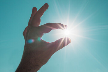 Hand of an adult man making an ok gesture over the blue sky and between his fingers catches the bright sun with its rays