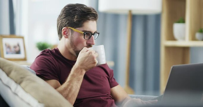 Male entrepreneur working online on a computer while drinking coffee sitting on a couch at home. Remote digital freelance worker typing an email and doing web research on a laptop indoors
