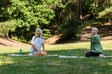 full length of senior woman pointing with finger near happy husband on fitness mat in green park.