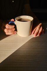 A cup of tea or coffee in the hands of a girl. In the morning, the girl drinks hot tea. Enjoy the comforts of home. White cup close-up in the hands of a woman in the rays of the sun from the window