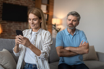 Dissatisfied adult caucasian husband looks at wife with smartphone, lady suffers from gadget addiction