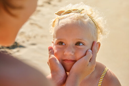 High Angle Of Cute Little Girl With Blonde Hair Looking At Mother Touching Face Tenderly On Sandy Beach On Sunny Day