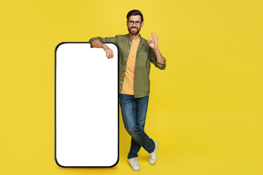 Positive stylish man showing okay gesture, standing next to huge smartphone with blank screen, advertisement mockup