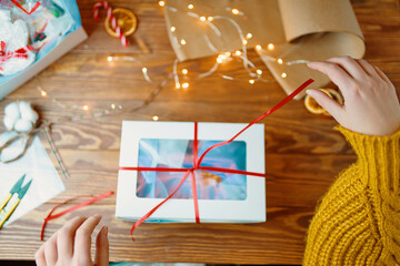 Flat lay of woman's hands in orange knitted sweater opening gift box. New Year's festive atmosphere. Christmas surprise with red ribbon and decorations on wooden table.