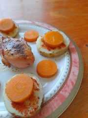 Plato gourmet de muslo de pollo con patatas en rodajas con condimentos y zanahorias decorado en círculo, forma un hermoso diseño culinario con luz suave con fondo de mesa de madera rústica