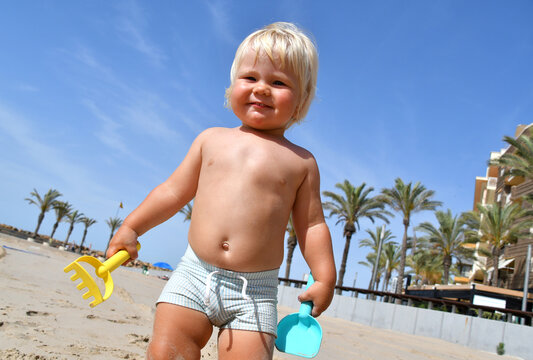 Portrait of a cute toddler boy playing with the sand at the beach in sunlight. Summer vacation with kids