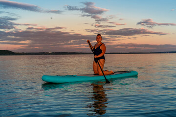 Fototapeta na wymiar a woman in a closed swimsuit on her knees on a SUP board with an oar floats on the water against the background of the sunset sky.