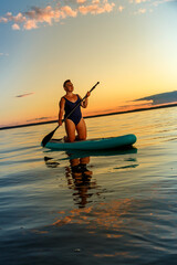 a Jewish feminist woman in a closed swimsuit with a mohawk on a SUP board with an oar floats on the water against the background of the sunset sky.
