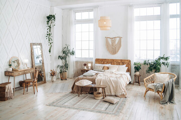 Fototapeta na wymiar Cozy rustic bedroom with boho ethnic decor. Bright spacious apartment with large windows. Wooden furniture. Boudoir table. Large mirror. Handmade textile. Plants in the interior. Empty space