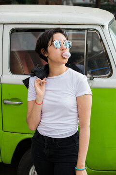 young greaser girl with short hair, posing with a fifties rockabilly style while chewing gum dressed in a white t-shirt black pants leather jacket and sunglasses with an old green hippie van (kombi) 