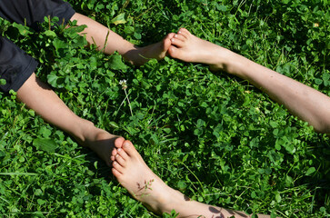 Naklejka na ściany i meble Small feet meeting bigger feet, feet contact of two kids sitting on the grass on a sunny bright day - concept of happy childhood, siblings sympathy.