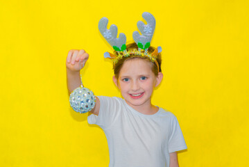 smiling boy with Christmas deer horns on his head holds a festive ball in his hand, stands on a...